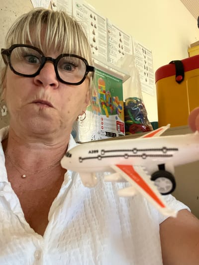 This is a close-up, horizontally oriented photograph of a woman holding a toy airplane. The woman has short blonde hair and is wearing large black-rimmed glasses. She is looking towards the camera with a neutral expression. A white collared shirt with a textured pattern is visible on her. A thin necklace with a small pendant is also visible.
In the background, a yellow and red object, possibly a storage container or toy box, is partially visible. Several sheets of paper with diagrams and text are spread out on a surface, possibly a table. A green box containing colorful building blocks is also in the background, along with a plastic bag filled with colorful rings. The overall setting appears to be an indoor space, likely a playroom or classroom. The visible text includes letters like 'G', 'H', 'R', and 'S', along with phrases like "by blocks will inspire hours of play and of builders" and "CE RECYCLEZ". The lighting suggests it is daytime, and there are no strong indicators of specific weather conditions or time of day.