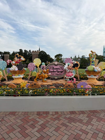 This is a medium shot, eye-level photograph of a whimsical display of Disney characters and oversized candy-themed decorations, likely situated at Tokyo Disneyland in Urayasu, Japan. The scene is a vibrant and colorful arrangement of exaggerated sweet treats and cartoon figures.

Dominating the center is a pink, ornate sign that reads "Sweets Garden Party" in elegant script. To the left of the sign, Mickey Mouse is depicted in a dynamic pose, standing atop a giant ice cream sundae overflowing with colorful whipped cream, macarons, and cookies. He is wearing a red and white outfit with a green scarf. Next to him is Pluto, the dog, in a playful stance, as if about to chase something.

To the right of the sign, Minnie Mouse is presented in a charming pink dress, holding a white rabbit. She is also positioned near a large ice cream sundae, similar in design to Mickey's. Further to the right, Goofy is standing, looking towards the viewer, with a waffle cone and assorted candies.

The entire display is set against a backdrop of green grass and trees, with a few distant structures resembling castles and modern buildings. The sky above is partially cloudy, with wispy white clouds against a pale blue expanse. The foreground features a neatly arranged flower bed with a variety of colorful flowers, predominantly yellow and orange, along with some pink and purple blooms. A dark metal railing separates the flower bed from the display. The ground in front of the flower bed is paved with reddish-brown bricks laid in a herringbone pattern.

The overall impression is one of a festive and cheerful outdoor environment, designed to evoke a sense of fantasy and fun, characteristic of a theme park.