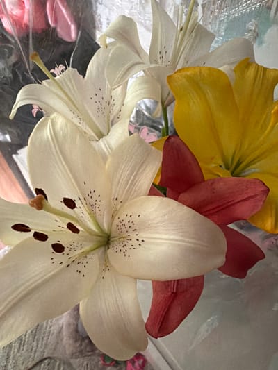 The image displays a close-up, overhead shot of a bouquet of lilies. The lilies are of various colors, including white, yellow, and red. Some of the white lilies have dark spots on their petals. The flowers are arranged in a way that suggests they are in a vase or wrapped for presentation. The background is blurred and appears to be indoors, possibly a home or a florist shop. There are no people or specific landmarks visible in the image. The image does not contain any information that would allow for the identification of its location in Volzhsky, Russia.