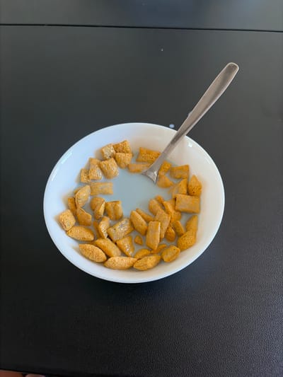 An overhead shot captures a white, round bowl filled with cereal and milk, positioned on a dark, textured surface, likely a table. The cereal consists of two main types: squarish, puffed pieces and more elongated, oval-shaped pieces, all appearing light brown or tan in color. Milk partially submerges the cereal, and a silver metal spoon rests within the bowl, its handle extending towards the upper left. The dark surface has a subtle granular texture, and a faint vertical line is visible on the left side, possibly an edge or seam of the surface. The scene depicts a simple meal, likely breakfast or a snack, within a domestic setting in Franklin Township, United States of America. No people are visible, and no text is discernible in the image. The time of day and weather are not evident.