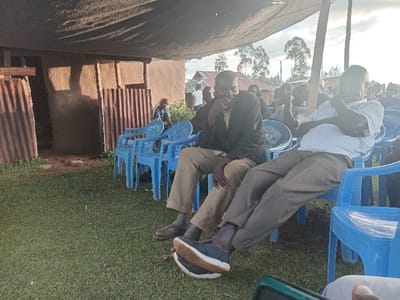 The image depicts an outdoor gathering in Chevaywa ward, Kenya, featuring several individuals seated on light blue plastic chairs on a grassy surface. A large, dark fabric canopy extends overhead, casting shadows. In the mid-ground, two adult males are prominent. The male on the left wears a dark blazer over a lighter shirt and khaki pants; he leans forward with his right hand near his chin, observing something to his right. The male on the right, dressed in a white short-sleeved shirt and light brown pants, leans back with his arms crossed over his chest, also looking right. To the left of the seated individuals, a mud-walled building with corrugated metal sheets leaning against its entrance or exterior is visible. Multiple additional people are present in the background, mostly seated on similar chairs, facing generally in the same direction. The distant background reveals trees and power lines under a bright sky. In the lower foreground, a hand holding a dark-colored electronic device is partially visible.