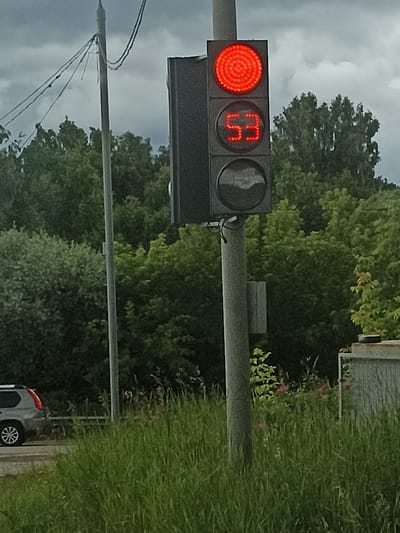 This media file shows a traffic light in Buturlino, Russia, on an overcast day. The traffic light is a modern, rectangular unit with three lenses. The top lens is illuminated with a bright red light, indicating a stop signal. The middle lens displays the number "53" in red LED digits. The bottom lens is dark and appears to be a pedestrian signal or another indicator.
The traffic light is mounted on a sturdy, grey pole. Behind the traffic light, dense green trees form a backdrop under a cloudy sky. To the left of the traffic light pole, a silver SUV is partially visible, driving on a road. The foreground is filled with tall, lush green grass. In the background, to the right, a portion of a building or structure with a textured grey facade is visible. The overall scene suggests an outdoor setting, possibly a road intersection or a highway. The weather appears to be cloudy, and the lighting suggests it is daytime, though the specific time is not evident. No people are visible in the image. The text visible is the number "53" on the traffic light.