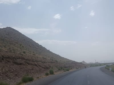 The media file depicts a scenic view of a road winding through a mountainous, arid landscape. The foreground features a paved road with white lane markings, stretching into the distance. To the left, a steep, rocky hillside is covered with sparse, dry vegetation and small shrubs. The hillside appears to be composed of various shades of brown and grey rock.

In the middle ground, the road curves to the right, leading towards a vast, open expanse. Several scattered, small, conical hills or mountains are visible in the distance, under a hazy, overcast sky. There are also utility poles with flags on them dotting the landscape. A single motorcyclist is seen riding on the road in the mid-distance, appearing as a small figure against the vastness of the terrain.

The sky is pale blue with scattered white clouds, suggesting it is daytime, likely late morning or early afternoon, given the soft light and lack of harsh shadows. The overall weather appears to be hazy, possibly due to dust or humidity, which softens the distant features. The scene evokes a sense of a remote, somewhat desolate, but beautiful natural environment. The location context points to Qilla Saifullah, Pakistan, which is consistent with the arid, mountainous terrain depicted. There is no visible text in the image.