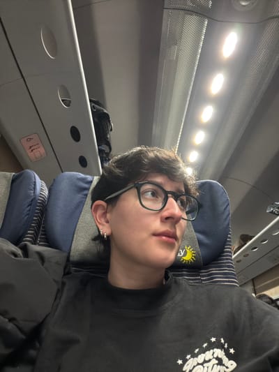 A young person with dark hair and round glasses reclines on a blue and gray striped headrest. They are wearing a black shirt with white graphics on the right side. The person is angled so that they are looking slightly upward and to the left. Above them is a perforated metal panel with a row of circular lights. To the left, a gray luggage rack with circular cutouts is visible, and a black bag is partially visible behind it. The scene appears to be inside a train or airplane.