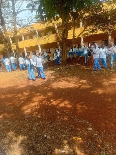 A group of people stand in front of a yellow building, possibly a school or institution. The individuals appear to be students, clad in light-colored shirts and blue pants. A tree with dense foliage frames the left side of the image. The ground is predominantly brown and earthy, with scattered debris visible. The picture's orientation is vertical.