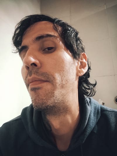 A low-angle, close-up headshot of a man with dark hair and a short beard. He is wearing a dark-colored hooded sweatshirt. The background appears to be a tiled wall, possibly in a bathroom or kitchen. The location is unspecified and cannot be determined from the image alone.