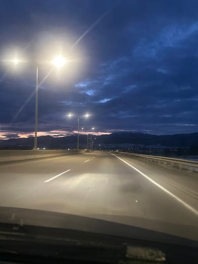 A highway at dusk stretches forward, reflecting light. Streetlights illuminate a dark blue, cloudy sky to the left. Mountains are faintly visible in the distance below the sky. The highway pavement displays blurred movement. A partial view of the vehicle's interior is on the right.