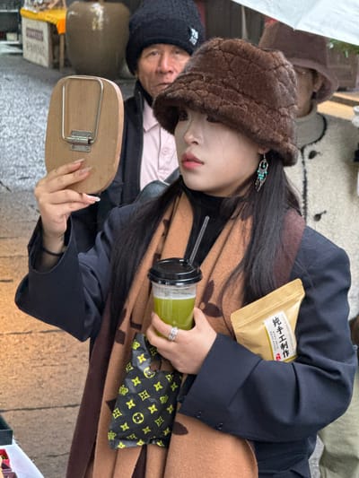 A young East Asian female is centrally depicted in an outdoor urban street or market environment within Pengshui Miao and Tujia Autonomous County, China. She wears a brown faux fur bucket hat, a dark high-collared top, a navy blue blazer, and a tan scarf featuring dark patterned monograms. Her right hand holds a small, rectangular wooden hand mirror with a metal stand. Her left hand grips a clear plastic cup containing a green liquid, sealed with a black lid and straw. Tucked under her left arm are a small black plastic bag with yellow repeating patterns and a light brown paper pouch marked with Chinese characters "纯手工制作" (Chún shǒugōng zhìzuò). She wears a silver-toned ring on her left ring finger and a turquoise and silver-toned earring on her left ear. Behind her to the left, an older East Asian male, wearing a black knit beanie, a pink shirt, and a dark jacket, is partially visible, observing the scene. To the far right in the background, another individual, mostly obscured, holds a white umbrella. The background also shows large cylindrical ceramic pots and a market stall with yellow fabric. The ground appears to be paved.