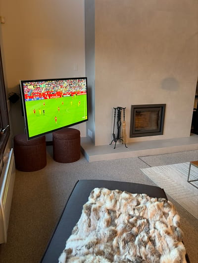 The image depicts an interior scene, likely a living room, possibly located in Mont-Tremblant, Canada, given the context. A large flat-screen television is mounted on the wall, displaying a soccer match. Below the television are two dark reddish-brown cylindrical ottomans. To the right of the television is a modern fireplace with a dark metal frame, set into a light-colored tiled wall. Fireplace tools, including a poker, tongs, and a brush, are neatly arranged on a stand next to the fireplace. A section of a grey couch is visible in the foreground, draped with a plush, multi-colored faux fur blanket. The flooring is carpeted. The environment appears to be a contemporary living space designed for relaxation and entertainment.