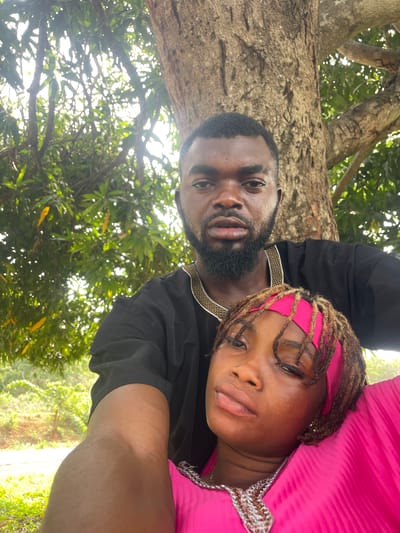 This image is a close-up selfie featuring two individuals, an adult male and an adult female, positioned horizontally against a natural outdoor backdrop in what appears to be Ogbe, Nigeria.
The male subject, situated on the left, has dark skin, a neatly trimmed beard, and short dark hair. He is wearing a black top adorned with an intricate gold and white patterned trim along the collar and shoulder line. His expression is neutral, and he looks directly towards the camera.
Beside him, the female subject also has dark skin and styled locs (dreadlocks), which are held back by a bright pink headband. She is dressed in a vibrant pink garment, possibly a top or dress, with some silver or white decorative elements near the neckline. Her eyes are partially closed, contributing to a relaxed or pensive demeanor as she also looks towards the camera.
The background is dominated by lush green foliage and the sturdy trunks of large trees, indicating a natural, possibly forested or park-like environment. Bright sunlight filters through the leaves, creating dappled light and suggesting a sunny daytime setting. No visible text is present in the image.