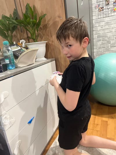A child in a black T-shirt and black shorts is seen from an elevated, oblique angle. He is kneeling on the floor near a light green exercise ball and holding playing cards. To his left, on a white surface, are various items including a plant, a white zippered bag, a calculator, bottles, and toiletries. A piece of blue tape is affixed to the white surface. The background includes a faux brick wall, wooden flooring, and part of a cabinet. There is no information to suggest this image was taken in the city of Vesyoloye, Russia.