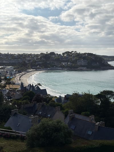 This is a photograph taken from an elevated viewpoint overlooking a coastal town and its beach. The scene is dominated by the sea, which is a pale turquoise color with small waves breaking on the shore. A crescent-shaped beach runs along the coastline, populated by a scattering of people and what appear to be beach umbrellas.
Behind the beach, the town is built on a steep hillside. Buildings, varying in size and architectural style, are densely packed, climbing up the incline. Many of the houses have dark tiled roofs. Lush green trees and vegetation are interspersed throughout the town and cover the slopes of the hill, creating a natural contrast with the built environment.
In the foreground, closer to the viewer, are more houses with dark, sloped roofs, some featuring dormer windows. A dense canopy of dark green trees is also prominent in this area, suggesting a wooded hillside.
The sky above is filled with dramatic, layered clouds. Some are bright white and illuminated by sunlight, while others are darker and more textured. The lighting suggests it is either early morning or late afternoon, with the sun casting long shadows and creating a soft glow. The weather appears to be partly cloudy, with a bright, possibly breezy day.
There is no visible text in the image. The overall impression is one of a picturesque, bustling coastal community in France, possibly during the summer season, given the presence of people on the beach.