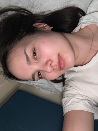 The scene takes place indoors in the city of Druzhinino, Russia. The image displays a close-up, angled perspective of an individual, identified as female, lying down. Her head is positioned towards the lower left of the frame, with her body extending towards the upper right. She possesses dark brown, long hair, which is spread across a light-colored, textured fabric surface, consistent with a pillow or bedsheet. Her eyes are open, dark brown, and directed slightly upwards. Her eyebrows are dark and well-defined, and her lips are slightly parted, revealing a natural pink hue. Her skin appears fair and generally unblemished. The individual is wearing a white short-sleeved t-shirt with a rounded neckline. A delicate gold-colored chain necklace is visible on her neck. Her left ear is adorned with a small silver-colored hoop earring in the helix and a small stud earring in the earlobe. A dark blue, textured surface is visible in the lower left foreground, while a beige wall or furniture component is partially visible in the upper left background. The ambient lighting appears dim. The individual is in a supine or reclined position, looking towards the point of capture.