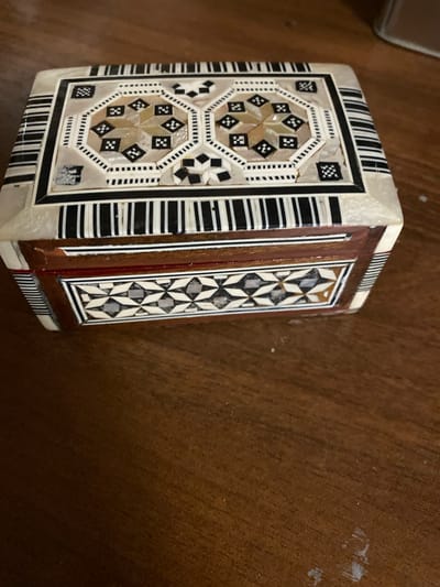 The image shows an ornate wooden box with intricate inlaid designs of geometric patterns and squares. The box has a light-colored top and sides with dark and light brown wood accents. The top of the box features an octagonal design surrounded by alternating black and white stripes. The sides of the box also have decorative inlay work. The box is placed on a dark brown wooden surface, possibly a table, with some light-colored dust or debris scattered on it. The background is out of focus, showing a blurry brown and beige area. The scene is a close-up shot, with the box filling most of the frame. There are no people or any indication of activity. There is no information in the image that suggests a location in Yokohama, Japan.