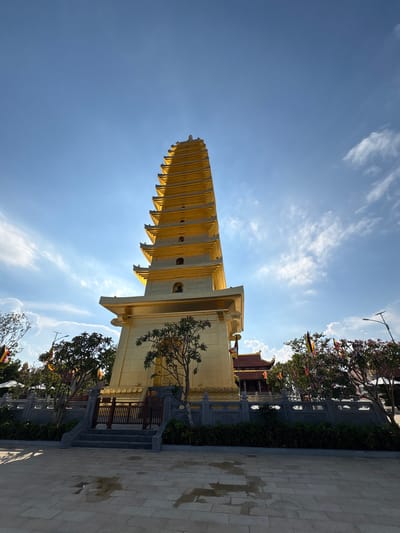 The image captures the Kim Son Pagoda, a prominent golden pagoda located in Diên Khánh, Vietnam. The pagoda's tiered structure reaches into a blue sky dotted with wispy clouds. A small tree grows near the base of the pagoda, next to a wooden railing and steps leading towards the structure. Beyond the pagoda, trees and decorative poles with colorful flags are visible. A paved area is in the foreground.