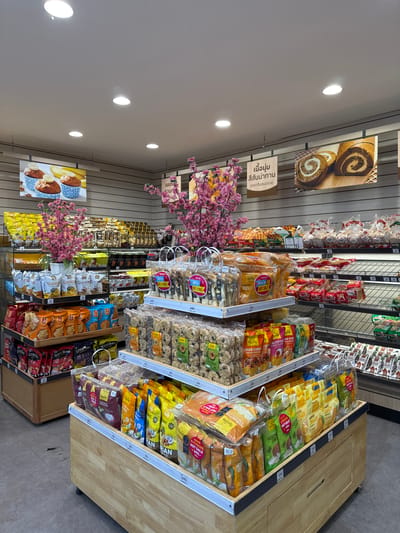 The image captures the interior of a grocery store in Thong Sala, Thailand. The perspective is from the aisle, focusing on shelves stocked with various packaged foods, including cookies, crackers, and jelly. Signage in Thai is visible, indicating product descriptions and promotional offers. Artificial cherry blossom decorations are interspersed throughout the displays, adding a decorative element to the retail environment. Fluorescent lights illuminate the scene from the ceiling.