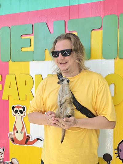 In the city of Phước Hải, Vietnam, a person with blond hair and sunglasses smiles while holding a meerkat. The person is wearing a yellow t-shirt with a black strap across their chest. The background features a colorful wall with large, cutout letters that appear to spell out "MENTION FARM". A cartoon meerkat sticker is visible on the lower left side of the wall. The scene suggests a visit to an animal-related attraction.