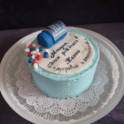 A light blue frosted cake is presented on a white plastic plate with a decorative paper doily. The cake is topped with a blue, pill-shaped decoration that has a silver label. Scattered around the pill decoration are smaller, edible pills in red, white, and blue. A white fondant circle on top of the cake features black handwritten Russian text that translates to "Happy Birthday, Masha! I wish you health and peace of mind." The background is dark, with the cake and plate positioned on a dark fabric surface. The overall scene is a close-up, eye-level shot of the cake. There is no discernible location in Aktobe, Kazakhstan.