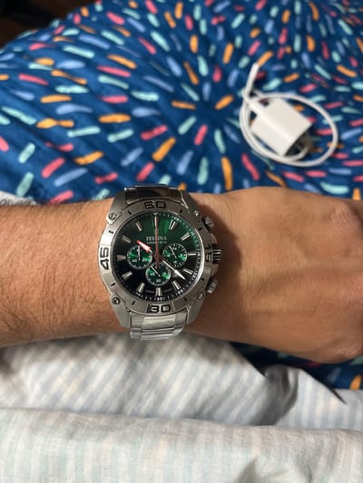 A wrist wearing a silver-tone Festina chronograph watch with a dark green dial is centered in the frame. The watch has a metal band and prominent bezel markings. To the left is a blue patterned textile. In the upper left, a white charger with a connected cable sits on the patterned textile. To the right is a light blue and white striped textile. The location and time are not discernible. The focus is on the watch and the arm on which it is worn.