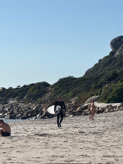 The image captures a section of Llandudno Beach in Hout Bay, South Africa, presenting a scene with diverse activities. At the edge of the beach near a rocky, vegetation-covered cliff, a man in a black wetsuit carries a white surfboard. Further up the beach, two individuals wearing bathing suits are standing. Towards the lower part of the frame, another man, shirtless and wearing dark swim trunks, is bending over. The beach surface appears textured with coarse sand. The sky is clear and bright blue.