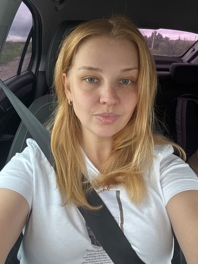 A blonde woman with fair skin and green eyes, wearing a white t-shirt and a seatbelt, is sitting in the driver's seat of a car. The image is taken from the chest up, with the woman looking directly at the camera. Her hair is shoulder-length and slightly wavy. There is a faint pattern of freckles on her nose and cheeks. She is wearing a small hoop earring in her left ear. The interior of the car is visible, including the dark headrest and part of the dashboard. The window to her left shows a blurry outdoor scene with a dirt road and vegetation. The overall lighting suggests it may be overcast or twilight. There is no visible indication of the location being in the city of Большое Село, Russia.