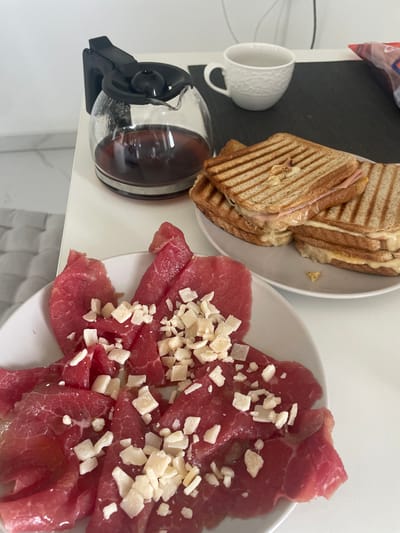 A high-angle shot displays a white table surface with a meal laid out, featuring sliced grilled cheese sandwiches on a white plate, a clear glass coffee pot filled with dark liquid, a small white cup, and a white plate with thinly sliced raw meat topped with small, irregular chunks of a light-colored substance, possibly cheese. The lighting is bright and appears to be natural. The scene is contained indoors, likely within a domestic setting. There are no people visible in the image, nor are there any indications of the location of the city of Kranj, Slovenia.
