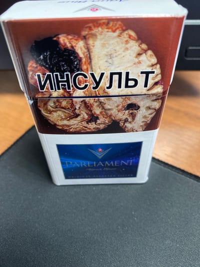 A close-up, eye-level, slightly angled shot shows a pack of Parliament Aqua Blue cigarettes. The pack is white with a dark red band across the top featuring an image of a brain with a dark mass on the left side. Bold black Cyrillic text that reads "ИНСУЛЬТ" (which translates to "stroke" in English) is superimposed over the brain image. Below this, the lower half of the pack is white with a blue rectangular emblem containing the "PARLIAMENT" logo and "Aqua Blue" text. The pack is placed on a dark grey textured surface, possibly a mousepad, with a wooden surface visible in the background to the right. There are no people or animals present in the image. The location is not discernable from the image itself.