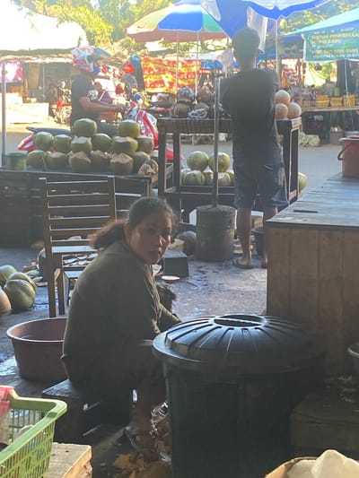 In Ta', Indonesia, an outdoor marketplace scene unfolds under a bright sun. A woman with dark hair, wearing a dark olive-colored shirt, sits on a low wooden stool. She is positioned in front of a tall, weathered wooden chair and to the left of a large black trash receptacle. Her gaze is directed towards the viewer. In the foreground, a red plastic basin is visible, and to the left, a green plastic basket rests on a wooden surface. The background reveals a bustling environment with various stalls. Several motorcycles are parked, and a person wearing a motorcycle helmet with a colorful design is seen. Stacks of coconuts are displayed for sale on tiered wooden tables. Further back, more stalls and merchandise are apparent, including what appears to be signage in Indonesian. The ground is paved and appears damp in places.