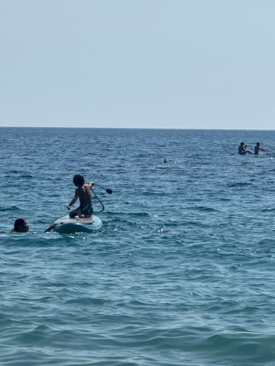 The image depicts a sunny day at Mirište, Montenegro, with a view of the Adriatic Sea. The primary subjects are several individuals engaged in water activities. In the foreground, a person is actively riding a wakeboard, appearing to be in mid-maneuver. Another individual's head is visible in the water nearby, suggesting they are swimming or also participating in water sports.
In the background, two figures are seen suspended in the air, connected by what appears to be a rope or harness, possibly engaging in a sport like parasailing or cable skiing. Further in the distance, a smaller dark object is visible on the water, its nature unclear.
The sea is a prominent feature, characterized by choppy, textured water reflecting the bright sunlight, creating shimmering highlights. The sky is a pale, clear blue, indicating fair weather conditions. The scene is set in a coastal environment, likely a popular beach or water sports area given the presence of multiple people and activities. There is no visible text in the image. The overall impression is one of active recreation and enjoyment on a beautiful summer day.
