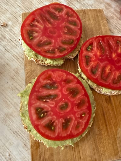 This is a close-up, overhead shot of three open-faced snacks on a wooden cutting board. Each snack consists of a round, puffed grain cracker topped with a light green spread, likely mashed avocado, and a thick slice of ripe red tomato. The tomatoes are cut crosswise, revealing their juicy interiors and seeds. The top snack is partially out of frame, while the other two are fully visible. The wooden cutting board has visible grain and scratch marks, suggesting it is well-used. The background surface is a light-colored, textured material, possibly a tablecloth or countertop. The lighting appears to be natural, casting soft shadows. The overall impression is one of a simple, healthy snack being prepared or served. No people are visible, and there is no visible text. The scene suggests a casual meal or snack time.