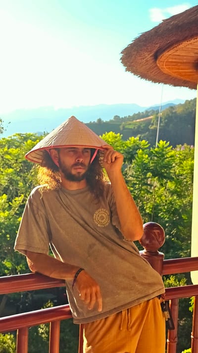 A man in his 20s or 30s is captured in a sunlit outdoor setting, leaning against a red wooden railing. He has long, dark, curly hair and a beard, and wears a traditional Vietnamese conical hat (nón lá) with a pink chin strap, which he is holding with his right hand. His gaze is directed thoughtfully towards the left. He is dressed in a faded greyish-green t-shirt featuring a circular graphic on the left chest area, which includes text like "RISE WITH THE SUN" and "SHINE WITH THE SUN" surrounding a sun or mandala-like design. He also wears mustard yellow or tan drawstring shorts, and a dark bracelet on his left wrist. A black lanyard or keychain with the text "GCS" and downward-pointing arrows is visible hanging from his shorts.
The scene is bathed in warm, bright sunlight, suggesting either early morning or late afternoon. The background is lush with vibrant green foliage, appearing as hills or cultivated terraces, and distant mountains under a clear, bright blue sky with some atmospheric haze. Part of a thatched roof structure is visible in the upper right corner, contributing to the rustic, traditional ambiance. The overall impression is one of serene observation amidst the scenic landscape of Mu Cang Chai, Vietnam.