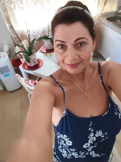 A woman with dark hair pulled back by a pearl-studded headband and wearing a dark blue dress with a white floral pattern poses for a selfie. She is wearing a gold necklace with a pendant and gold hoop earrings. She is seated in a room that appears to be a kitchen or dining area, with a white table and chairs upholstered in a floral fabric. A white air conditioning unit is visible to the left. A potted orchid sits on the table. The background includes a sink and cabinets, suggesting the location is likely an apartment in Livorno, Italy. The lighting is bright, suggesting daytime.