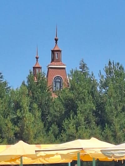 The image shows a bright, sunny day in Khudat, Azerbaijan. In the background, two ornate, copper-colored towers with spires rise above lush green trees. The towers have arched windows, and one appears to have a patterned roof. In the foreground, the tops of several yellow awnings are visible, suggesting an outdoor market or gathering. The sky is a clear, vibrant blue.