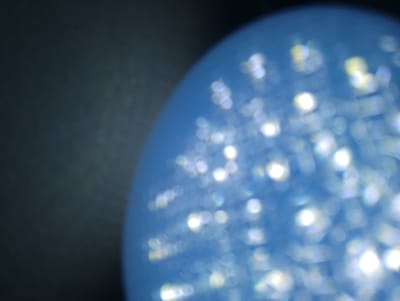 The image depicts a close-up, abstract view of a blue, spherical object with a dark, textured background on the left side. The blue surface is illuminated, creating numerous bright, out-of-focus circular highlights that vary in size and intensity. Some highlights appear white and sharp, while others are softer and tinged with yellow or blue. The overall impression is one of a shimmering or bubbly surface. There are no visible people, identifiable objects, or any contextual clues that would allow for a determination of its location within Katon Rikkos, Nigeria, or any other specific geographical setting.