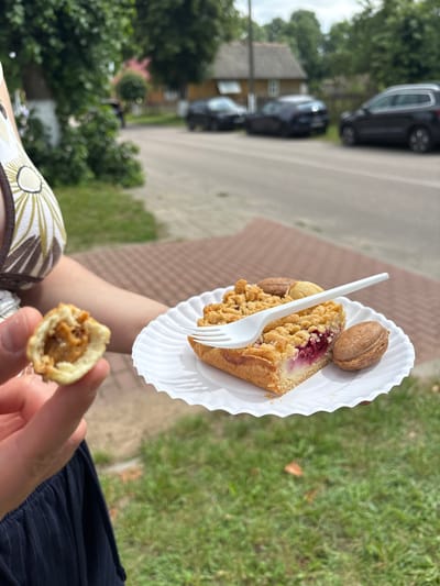 In Trześcianka, Poland, on a cloudy day, a person holds a half-eaten pastry in their left hand, revealing a caramel-like filling. In the background, a white paper plate holds a slice of fruit crumble cake with berries and a streusel topping. A white plastic fork rests on the cake. Beside the cake are two nut-shaped cookies. The plate is on a patterned red brick surface, with a grassy area to the right. In the further background, a street is visible with several parked cars and houses.