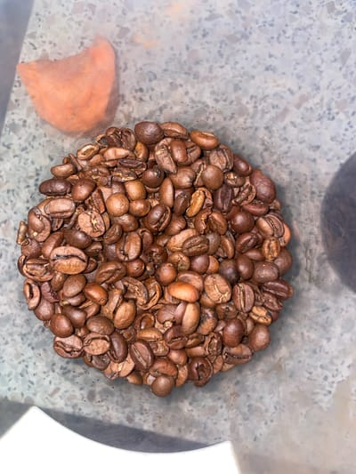 A close-up, overhead view shows a pile of roasted coffee beans. The beans are various shades of brown, with some appearing darker and more roasted than others. They are irregularly shaped, with a visible crack down the center of most beans. The coffee beans are arranged in a circular mound on a speckled, gray surface that appears to be a countertop. In the upper left corner of the image, a translucent, orange object is partially visible. This object is amorphous and appears to be a piece of food, possibly a dried apricot or a piece of sweet potato. The lighting is dim, casting subtle shadows on the beans. The overall impression is one of abundance and a focus on the texture and color of the coffee beans. There is no indication of the specific location within Laishevo, Russia.
