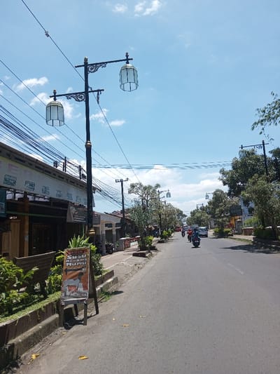 The image depicts a sunlit street in what is identified as Tagtse Tashidingkha, Bhutan. However, a prominent A-frame sign on the left side of the road displays the Indonesian text 'SEDIA Susu Kambing Humi.' The scene looks down a paved asphalt road under a clear sky with scattered clouds. Decorative black lampposts, each with twin lantern-style fixtures, line both sides of the street, interspersed with trees and green shrubs along the sidewalks. The left side features a row of single or two-story buildings with dense utility wires strung overhead. Multiple motorcycles with riders and at least one car are observed traveling on the road away from the viewer.