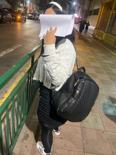 A woman stands on a sidewalk at night in Iquique, Chile, holding a piece of paper in front of her face. She is wearing a white cable-knit sweater, a black skirt, black leggings, and white sneakers with black stripes. A black backpack with gold zippers is slung over her shoulder. The sidewalk is paved with square tiles. A green metal railing runs along the left side of the frame, separating the sidewalk from a street. Cars are visible in the background, and two men are running in the distance. The streetlights cast a warm glow on the scene.