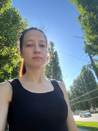 A woman is taking a selfie outdoors. She is wearing a black tank top and her hair is pulled back in a ponytail. She has a neutral expression. The background shows a clear blue sky, green trees, power lines, and a street with a car driving by. The location is the city of Voronezh, Russia.
