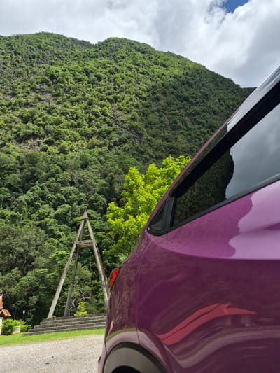 A purple car is parked on a gravel surface in front of a steep, lush green hillside. The hillside is densely covered with trees and vegetation, with some rocky outcrops visible. In the middle ground, there is a concrete structure that resembles an A-frame or a gateway, with a series of concrete steps leading up to it. To the left of the steps, there is a small building with an orange roof and white walls, and a lamppost. The sky is cloudy with some patches of blue. The reflection of a red-roofed building can be seen on the car's surface. This scene is located in Phang-nga, Thailand.