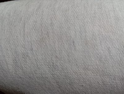The image is a close-up, abstract shot of a textured, light-colored fabric, likely cotton or a blend. The fabric appears to be a light grey or off-white, with subtle variations in shading and a visible woven pattern of interlocking threads. There are a few faint, darker smudges or stains scattered across the surface, particularly in the central and upper-left portions of the image. The lighting is soft and diffuse, highlighting the texture without creating harsh shadows. There are no identifiable objects, people, or environmental features present; the image is solely focused on the fabric's surface. The provided crops offer slightly different perspectives and degrees of focus on this same fabric. The image does not provide any information or context related to the city of Hlynytsi, Ukraine, or any specific location within it.