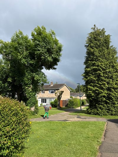 The image depicts a residential street scene in Hazlemere, United Kingdom, under a partly cloudy sky with a faint rainbow visible in the distance. A child, wearing bright green trousers and a hooded jacket, walks away from the camera carrying a large, colorful umbrella. The child is on a paved pathway in front of a brick house with a white front door and a bay window. To the left of the house is a large, mature tree with lush green foliage, and to the right, a tall evergreen tree. A neatly trimmed hedge borders the foreground on the left, and a patch of green lawn with scattered yellow flowers is visible. Another paved path curves towards the right side of the frame, and more houses and trees are visible further down the street. The overall impression is of a suburban environment.