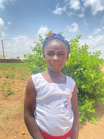 The image depicts a young girl in Turu, Nigeria, standing outdoors on a sunny day. She is wearing a white top with a patch on the chest, a red skirt, and a blue headscarf. Behind her is a green bush and a field of sparse vegetation. Power lines are visible in the background. The ground is a mixture of brown soil and grass. The sky is blue with scattered white clouds.