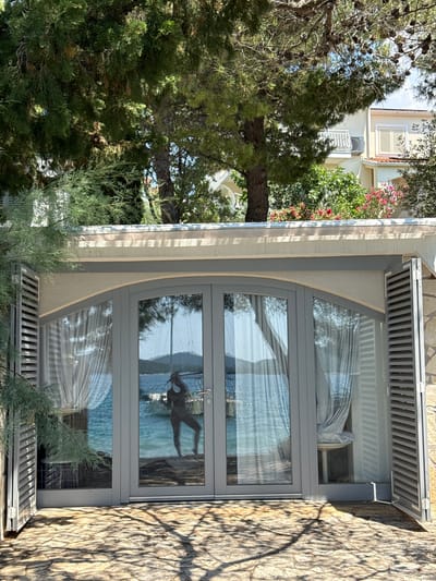 A full outdoor shot captures a reflective glass-door entrance to a structure in Grad Šibenik, Croatia. The entrance features a gray arched frame with two glass doors, flanked by gray louvered shutters. The doors reflect a serene scene of a bright blue sea with a boat and a figure standing on a shoreline. Behind the entrance, a white building with yellow accents and pink flowering bushes is visible through the foliage of pine trees. The ground in front of the entrance is paved with irregular stone slabs, shaded by dappled sunlight filtering through the trees. The overall scene evokes a tranquil coastal atmosphere.