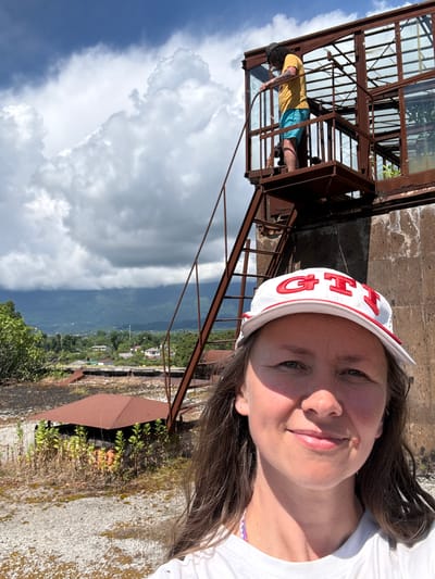 A woman wearing a white cap with "GTI" in red letters and a white t-shirt poses for a selfie outdoors. Behind her, on a rusted metal structure with glass windows, a person wearing a yellow t-shirt and blue shorts stands on a platform. In the background, a lush green landscape with mountains under a cloudy sky is visible. The scene appears to be in a somewhat dilapidated industrial or historical site, possibly in the city of Unknown, Abkhazia.
