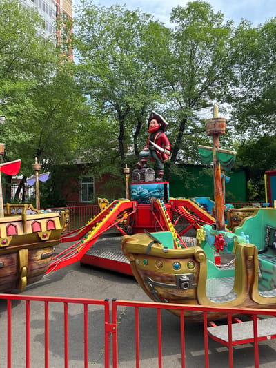 This is a vertical, eye-level, medium shot of a pirate-themed amusement park ride in Abakan, Russia. The ride features a ship with colorful seats and decorative elements like pirate figures and barrels. A red metal ladder is visible on the right side of the frame, suggesting access to the ride. The ride is stationary, and no people are actively using it, implying it is either not in operation or currently between cycles.

The setting appears to be an outdoor amusement park or fairground. Lush green trees with dense foliage surround the ride, providing shade and a natural backdrop. In the background, parts of buildings and a fence can be seen, indicating an urban or suburban location. The lighting suggests it is daytime, likely a bright day given the shadows. The overall atmosphere is cheerful and playful, characteristic of an amusement park environment. There is no visible text in the image.