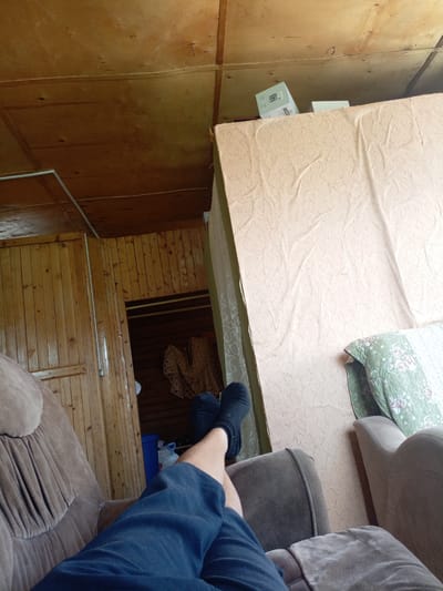 The image depicts an interior scene, likely within a residential dwelling in Churapcha, Russia. In the foreground, a person's legs and feet are visible, resting with one leg extended and the other bent, wearing dark socks and navy blue pants. The person is seated in a plush brown armchair. To the right, another armchair is partially visible. Behind the armchair, a tall, light-colored mattress leans against a wall covered in a textured, light-peach fabric. Above the mattress and continuing to the left is a wooden ceiling. To the left of the frame, a wall paneled with light-colored wood features a doorway leading to a darker interior space where a blue bin and a large white jug are visible. A lacy, cream-colored garment hangs within the doorway. The overall environment appears somewhat rustic and appears to be a living area.