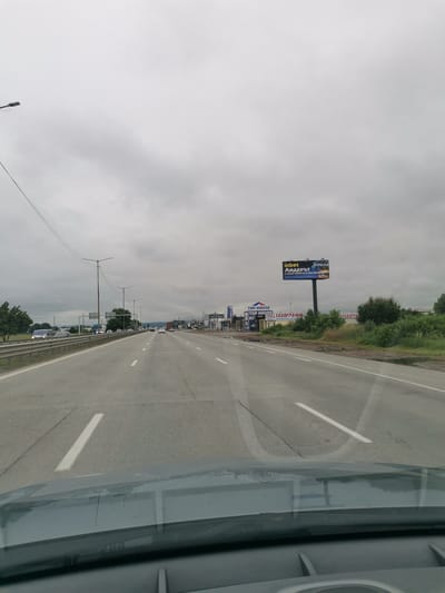 The image shows a highway in Kazichene, Bulgaria. The road is paved with asphalt and has white lane markings. There are several cars on the road, and a billboard advertising "inbet Лидерът" is visible on the right side of the road. The sky is overcast. The image is taken from inside a vehicle, as the top of the dashboard and windshield are visible in the foreground.