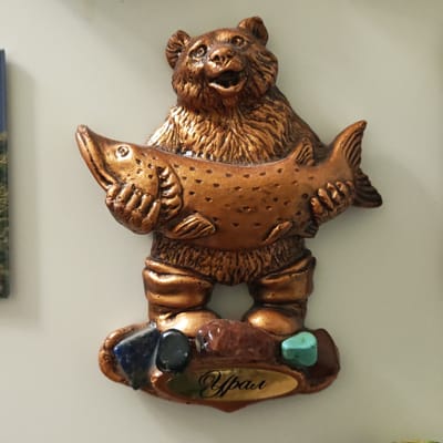 This image captures a close-up of a metallic-looking souvenir or magnet, likely from Novo-Nikol'skoe, Russia. The central subject is a bear figure, cast in a bronze or copper tone, standing upright and holding a large fish horizontally across its body with both paws. The bear has an open mouth, giving it an expression of joy or perhaps a roar, and features detailed fur texture. The fish also displays textured scales or spots.

The base of the figurine is decorated with several small, polished stones of various colors, including deep blue, black, reddish-brown, and turquoise, suggesting mineral wealth. Embedded in the base is a gold-colored oval plaque with the word "Урал" (Ural) written in elegant Cyrillic script, indicating the region this souvenir represents. The object is presented against a plain, light-colored background, characteristic of an indoor setting, possibly on a refrigerator or wall. The overall appearance suggests a tribute to the wildlife and natural resources of the Ural Mountains.