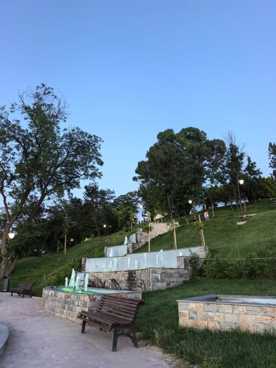The image depicts a terraced park area with a cascading fountain. The fountain features multiple levels with water jets, some illuminated with green light. Stone walls and pathways define the various levels. Two wooden park benches are visible on the paved area in the foreground. Lush green grass covers the sloping terrain. Several trees of varying sizes are scattered throughout the scene, with a prominent large tree on the left. The sky is clear and blue, suggesting it is daytime. The park appears to be located in Svishtov, Bulgaria, based on the provided context.